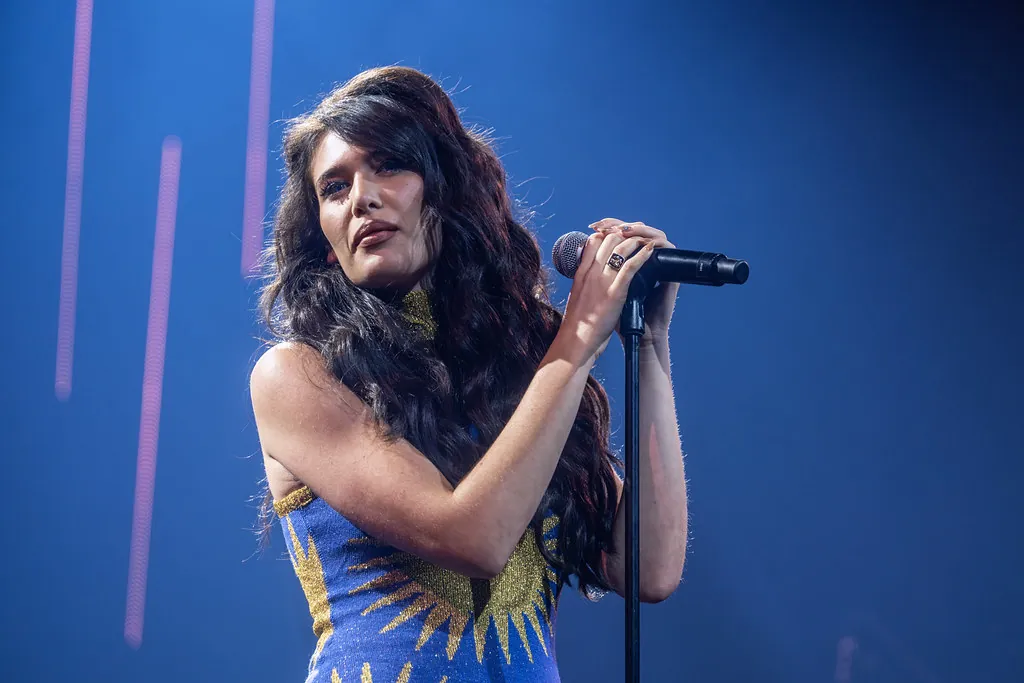 Sienna Spiro holding a microphone stand on stage in a blue and gold dress